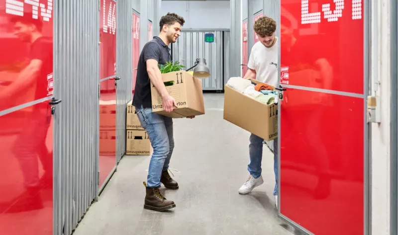 Bild von zwei Personen dien Kisten in einen Lagerraum stellen.