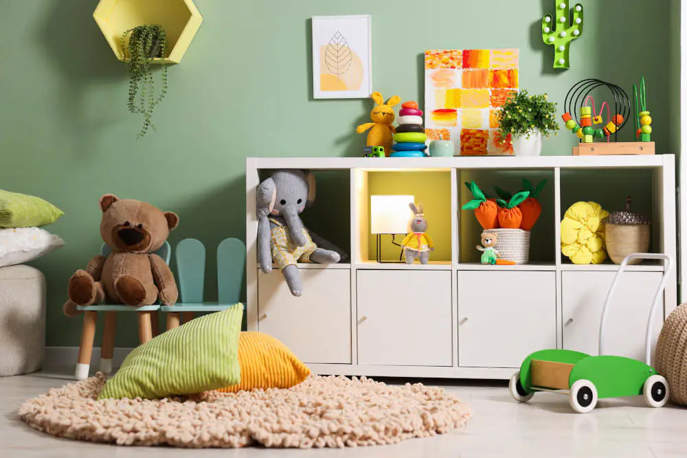 Einrichtung Kinderzimmer: Das Bild zeigt ein mit Spielsachen gefülltes Regal vor einer grünen Wand, auf einem Stuhl sitzt ein Teddybär, in dem Regal ein Elefant. An der Wand hängt eine Lampe in Kaktusform.