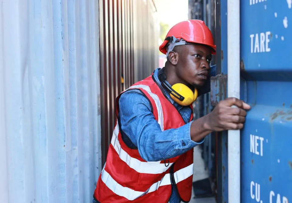 Vorteile und Nachteile von Lagercontainern. Ein Lagervorarbeiter mit Warnweste, Helm und Lärmschutz-Kopfhörern um den Hals öffnet einen Container, um hineinzuschauen