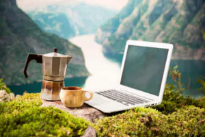 Ein aufgeklappter Laptop, ein Espressokocher und eine Kaffeetasse stehen auf einem bemoosten Felsabsatz vor einer atemberaubenden Gebirgslandschaft - als Sinnbild für eine Workation, die Arbeit und Urlaub verbindet.