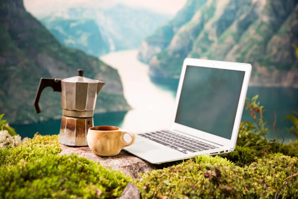 Ein aufgeklappter Laptop, ein Espressokocher und eine Kaffeetasse stehen auf einem bemoosten Felsabsatz vor einer atemberaubenden Gebirgslandschaft - als Sinnbild für eine Workation, die Arbeit und Urlaub verbindet.