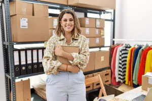 Eine junge Frau steht in ihrem E-Commerce-Lager, sie hält eine einen kleinen Karton im Arm, hinter ihr sieht man weitere Kartons sowie Aktenordner in einem Regal und eine Kleiderstange, an der Oberteile in verschiedenen Farben hängen.