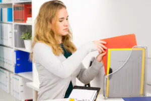 Produktivität im Büro steigern: Eine junge blonde Frau sortiert Dokumentenmappen ein einem hellen Büro, das wie eine Anwalt-, Notar- oder Steuerkanzlei wirkt. Im Hintergrund sieht man ein Regal voller Ordner und Bücher.